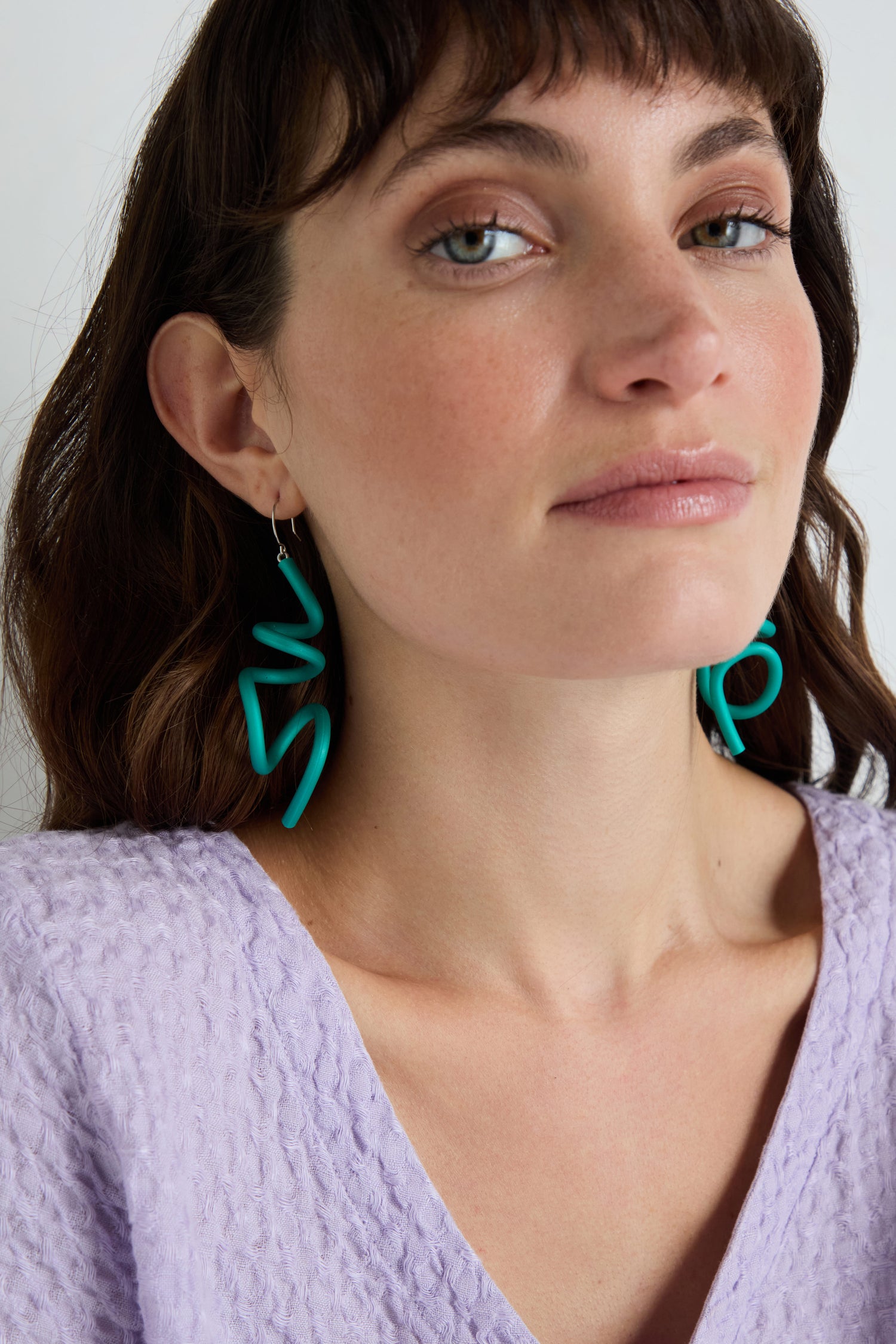 A woman in a light purple textured top wears large, sculptural Sketch Earrings in teal and looks calmly at the camera.