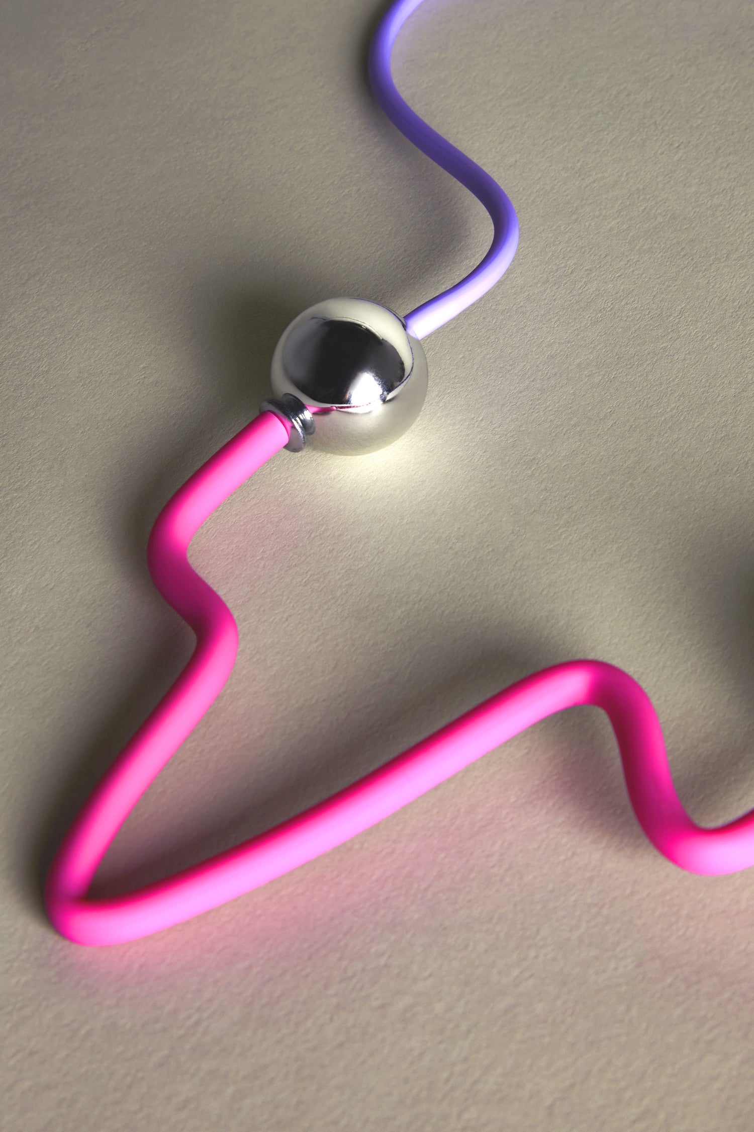 The Silver Sphere Colour Pop Necklace features a silver sphere connected to two flexible tubes—one pink and one purple—resting elegantly on a textured beige surface, echoing Samuel Coraux's unique style.