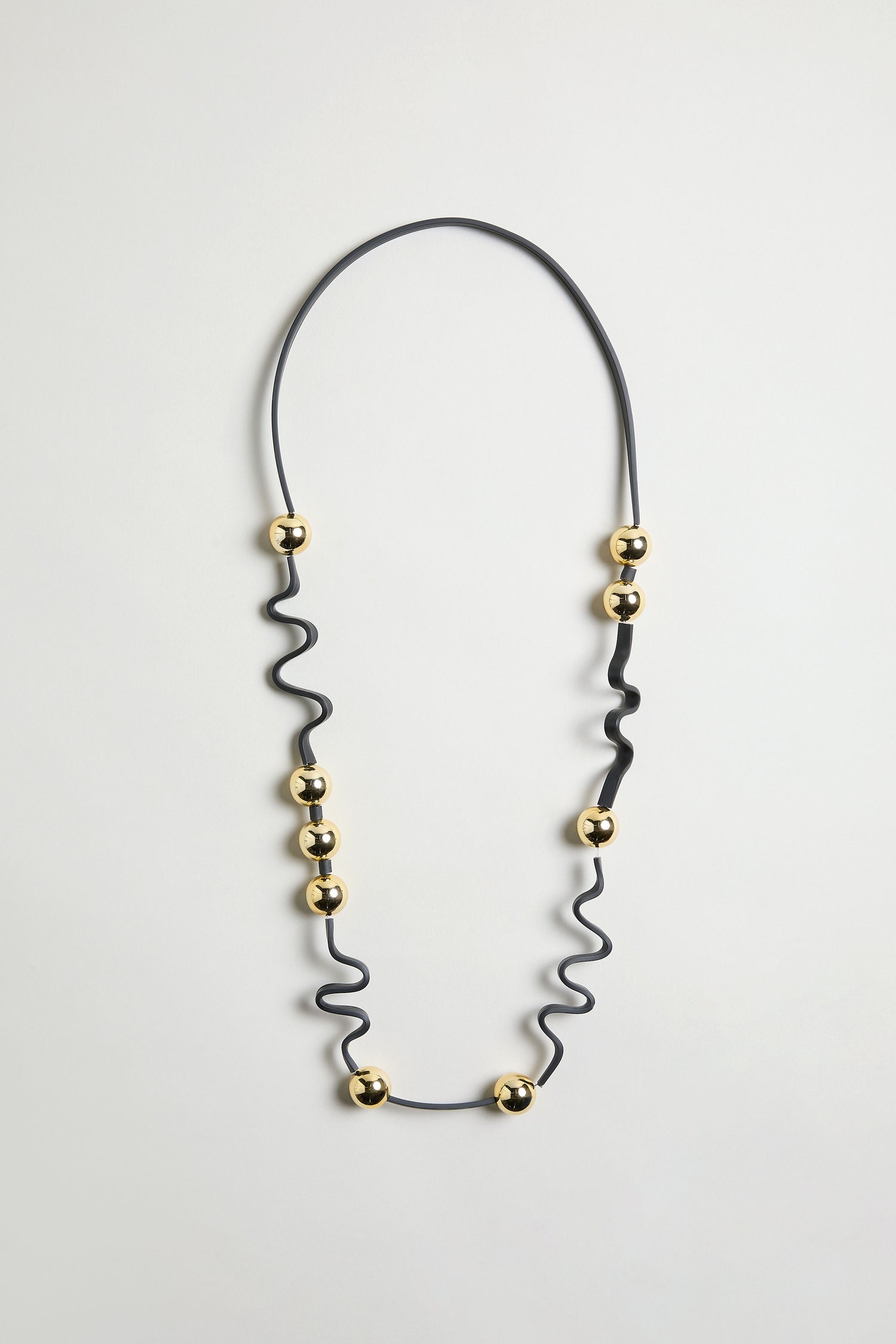 The Squiggle and Sphere Necklace is a black and gold piece with evenly spaced gold beads, forming a sculptural design that's showcased against a plain white background.