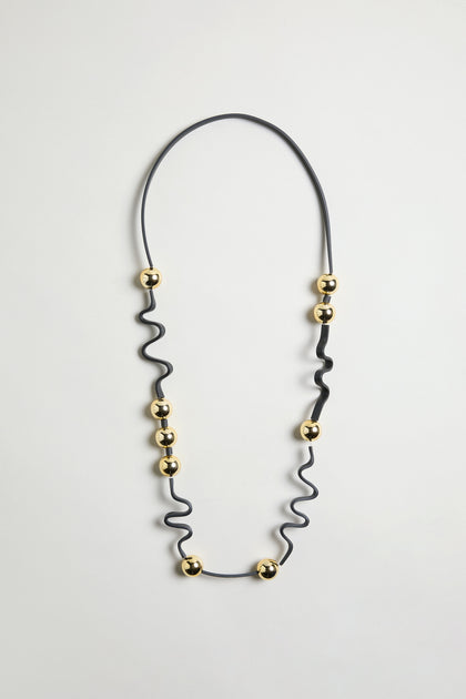 The Squiggle and Sphere Necklace is a black and gold piece with evenly spaced gold beads, forming a sculptural design that's showcased against a plain white background.