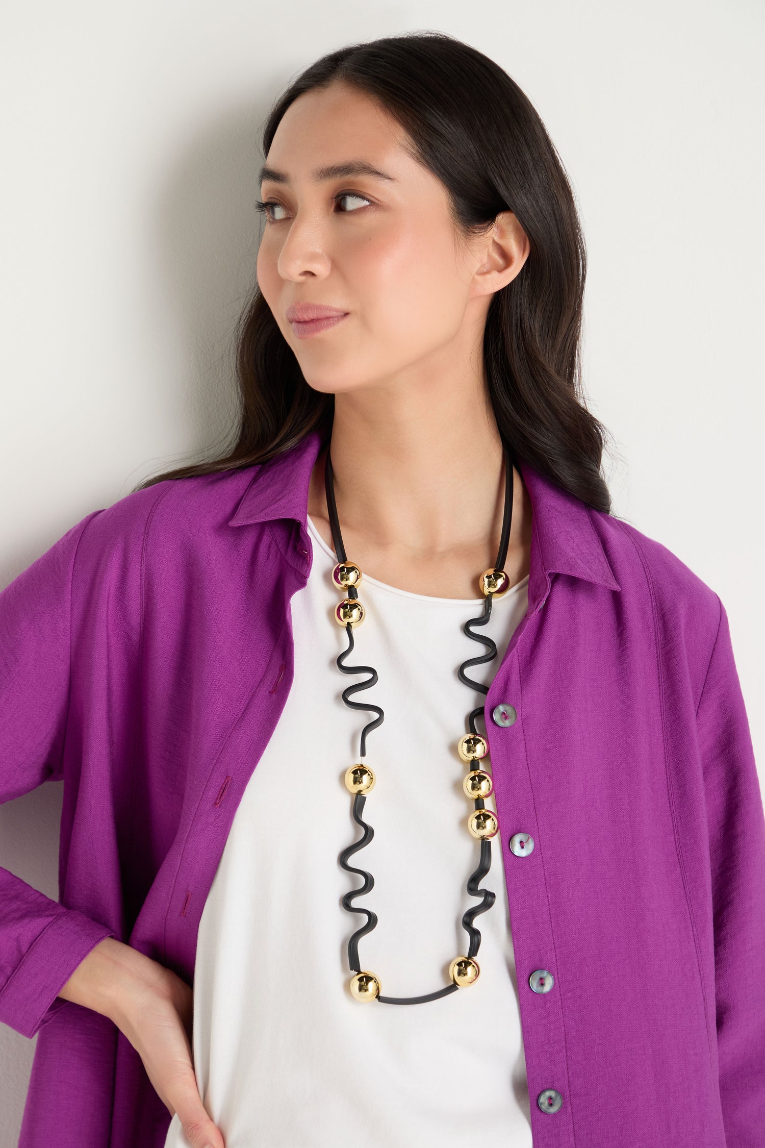 A woman stands against a plain light-colored wall, wearing a bright purple button-up shirt over a white top and the Squiggle and Sphere Necklace, a striking sculptural piece in black and gold.