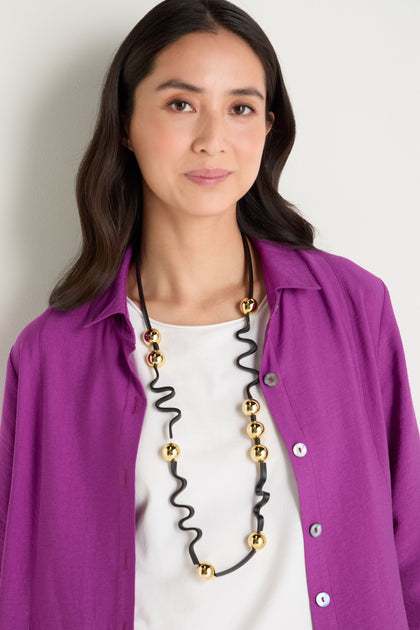 Woman with long dark hair wears a white top, bright purple open shirt, and the Squiggle and Sphere Necklace—featuring black and gold sculptural elements with gold beads—while standing against a light-colored wall.