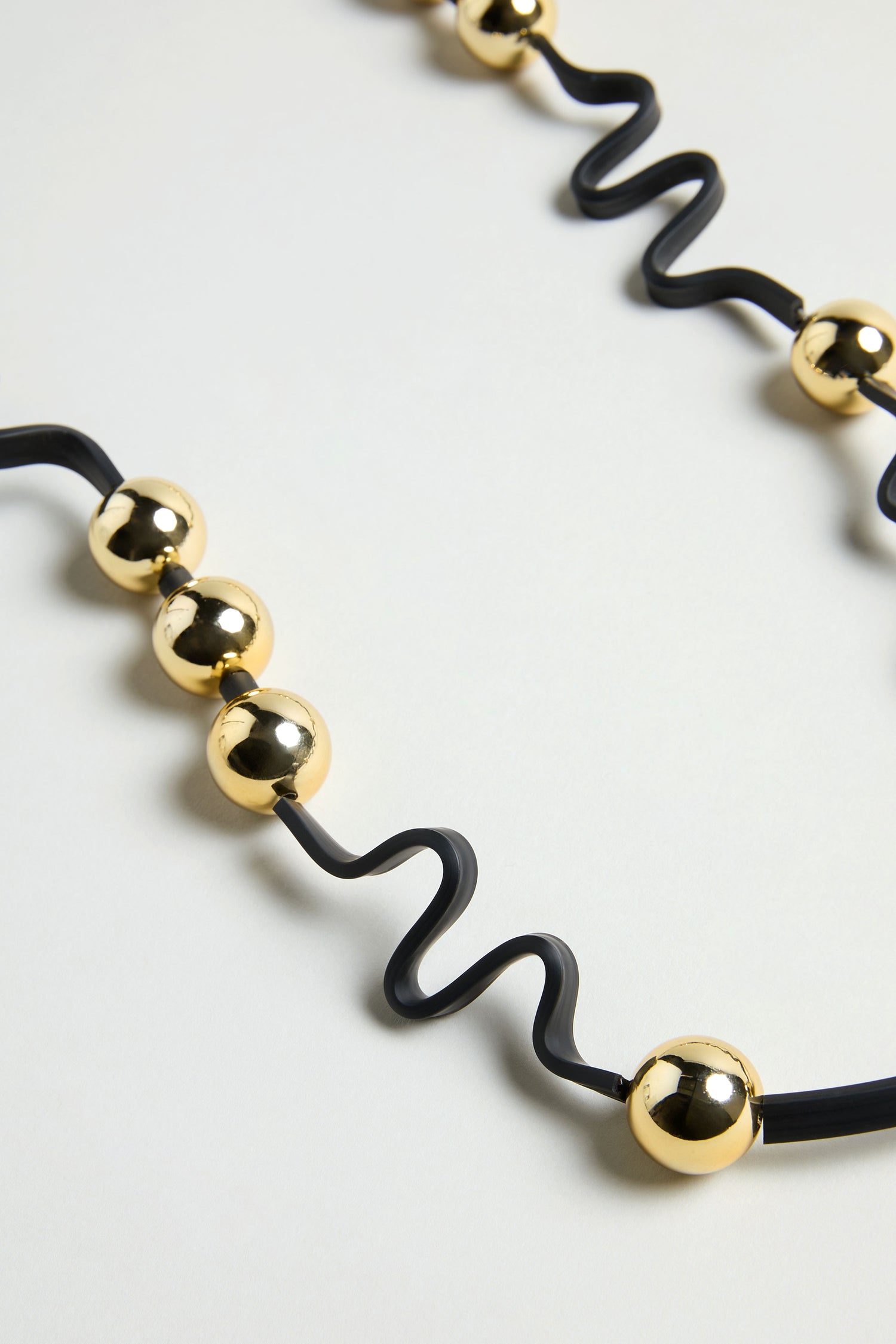 The Squiggle and Sphere Necklace features a sculptural black squiggle design accented with shiny gold spherical beads spaced along its length, displayed on a light surface.