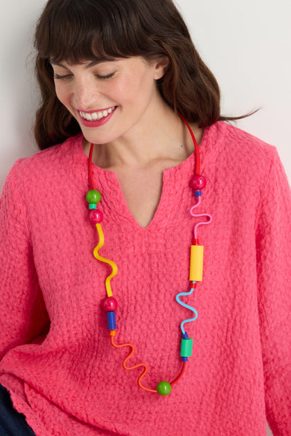A woman in a bright pink textured top looks down and smiles, showcasing the Rainbow Graphic Pop Necklace—an eye-catching piece featuring colorful wavy and cylindrical shapes that add a vibrant, sculptural touch to her look.