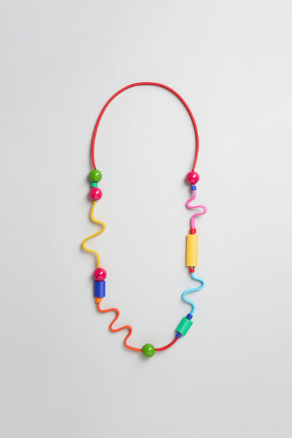 The Rainbow Graphic Pop Necklace is a sculptural piece with a red flexible band, colorful wavy segments, and large green, yellow, pink, and blue beads—handmade in Paris and shown on a plain light background.
