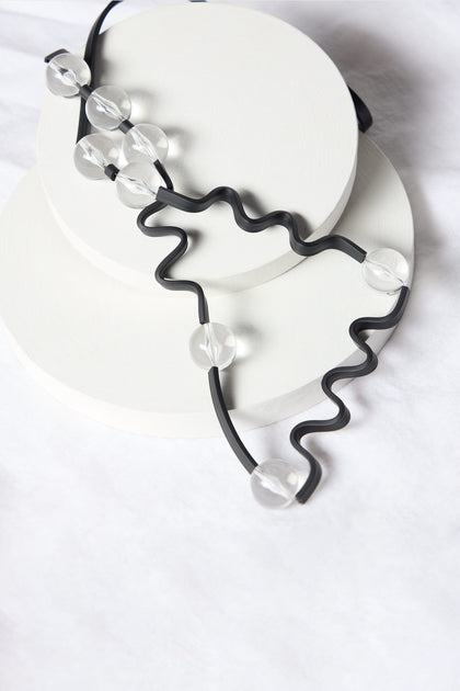 A Squiggle And Glass Necklace, boasting black wavy lines interspersed with clear beads, is elegantly showcased on two stacked white circular bases, capturing the distinctive style of Samuel Coraux.