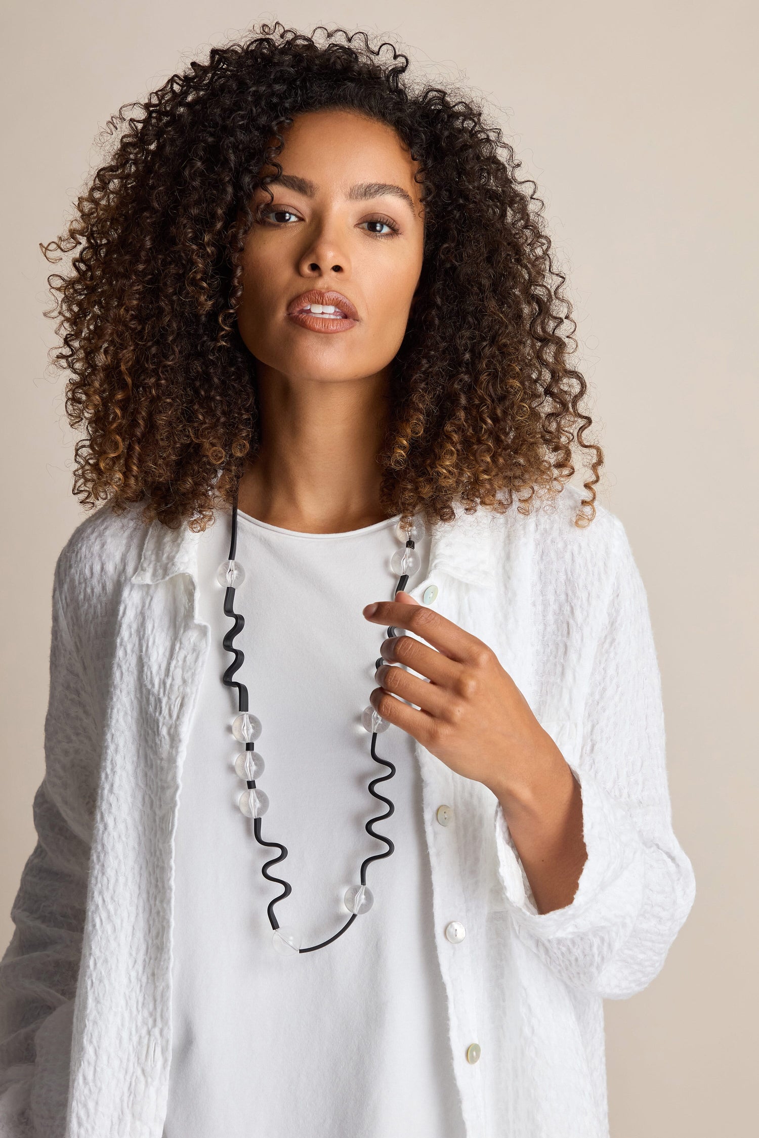 A person with curly hair wearing a white textured jacket poses against a plain background, holding the Squiggle And Glass Necklace adorned with Samuel Coraux designs.