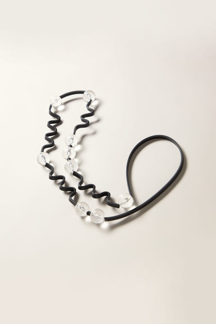 A Squiggle And Glass Necklace featuring a black elastic band adorned with delicately attached colored glass beads, arranged in an artful zigzag pattern against a light background.