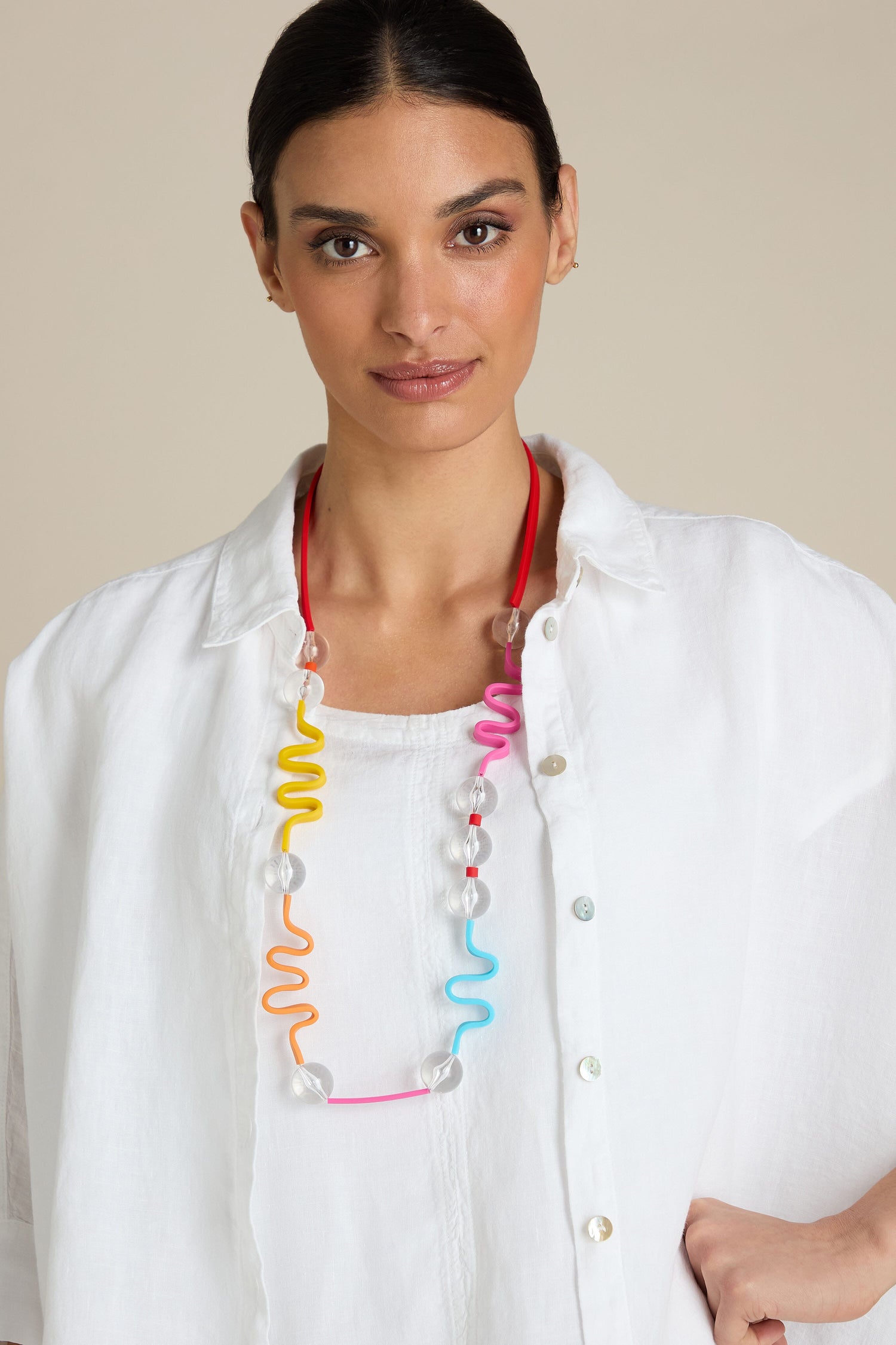 A person wearing a white shirt and the Squiggle And Glass Necklace, featuring colorful, translucent beads arranged in a unique abstract squiggle design, looks directly at the camera against a beige background.