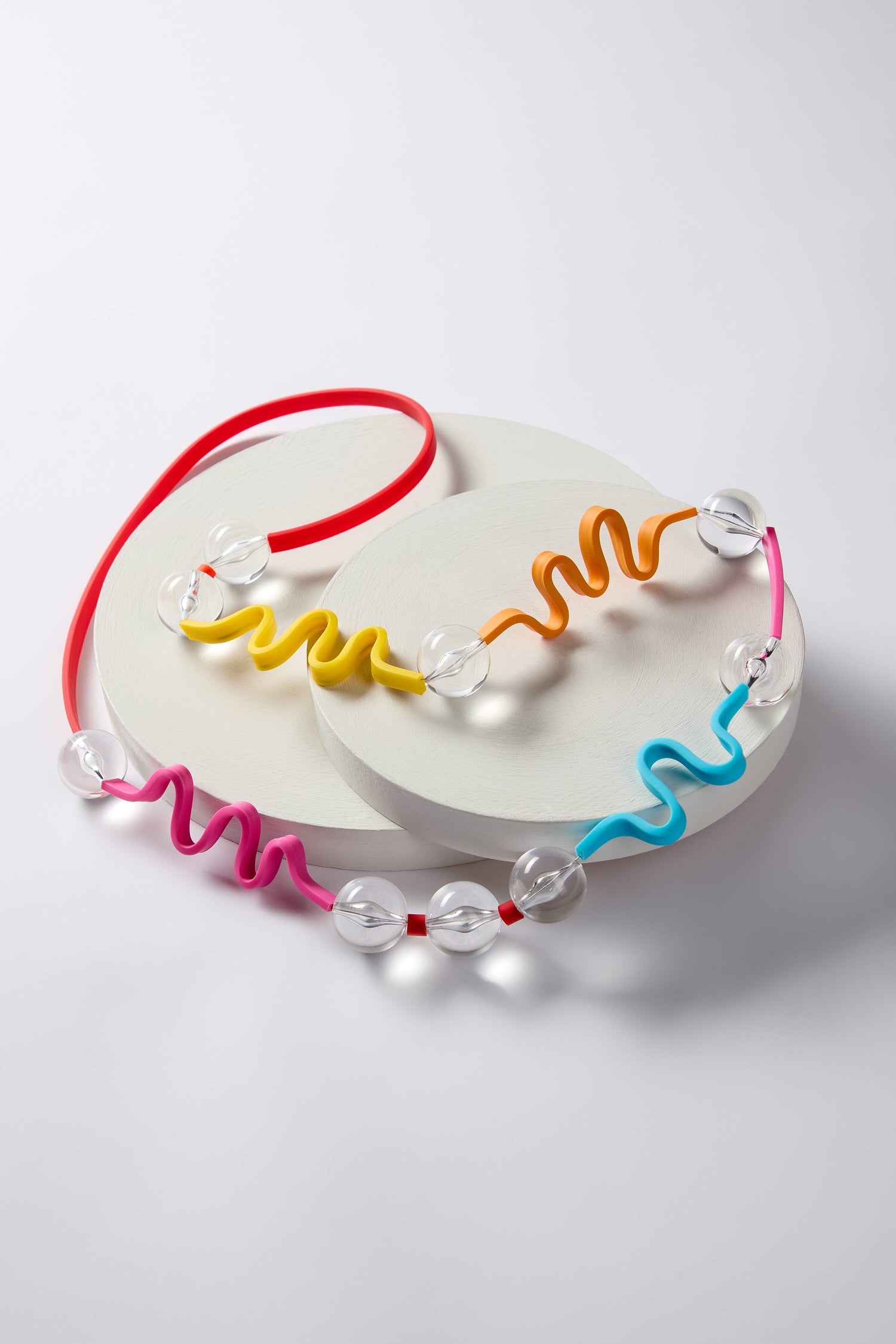 The Squiggle And Glass Necklace is a long-length accessory highlighted by its distinct abstract squiggle cord segments in red, orange, yellow, pink, and blue, interspersed with contrasting colored glass beads and elegantly displayed on white circular platforms.