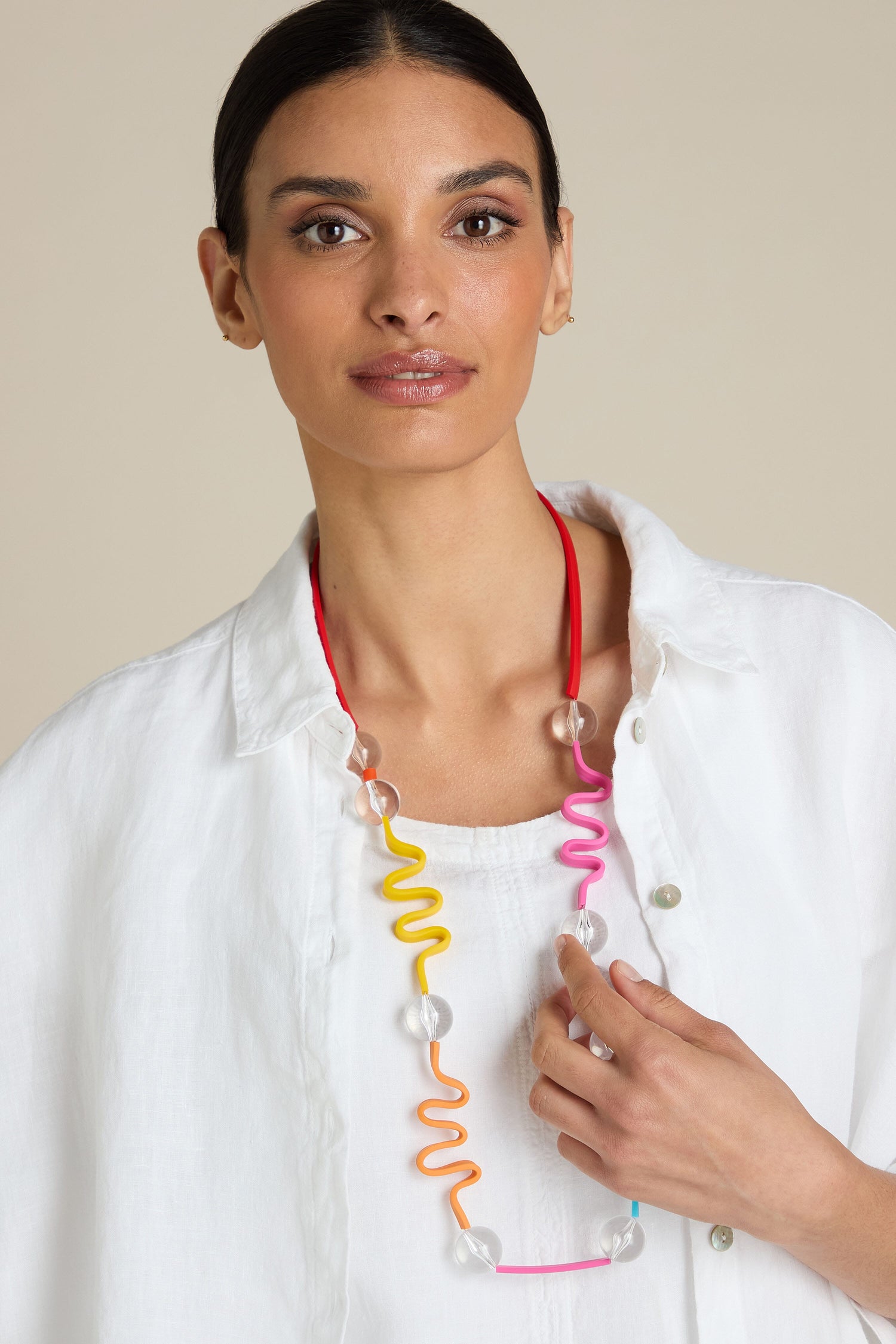 A person with dark hair is wearing a white shirt and the Squiggle And Glass Necklace, which features colorful, wavy designs with contrasting colored glass beads, while looking directly at the camera.