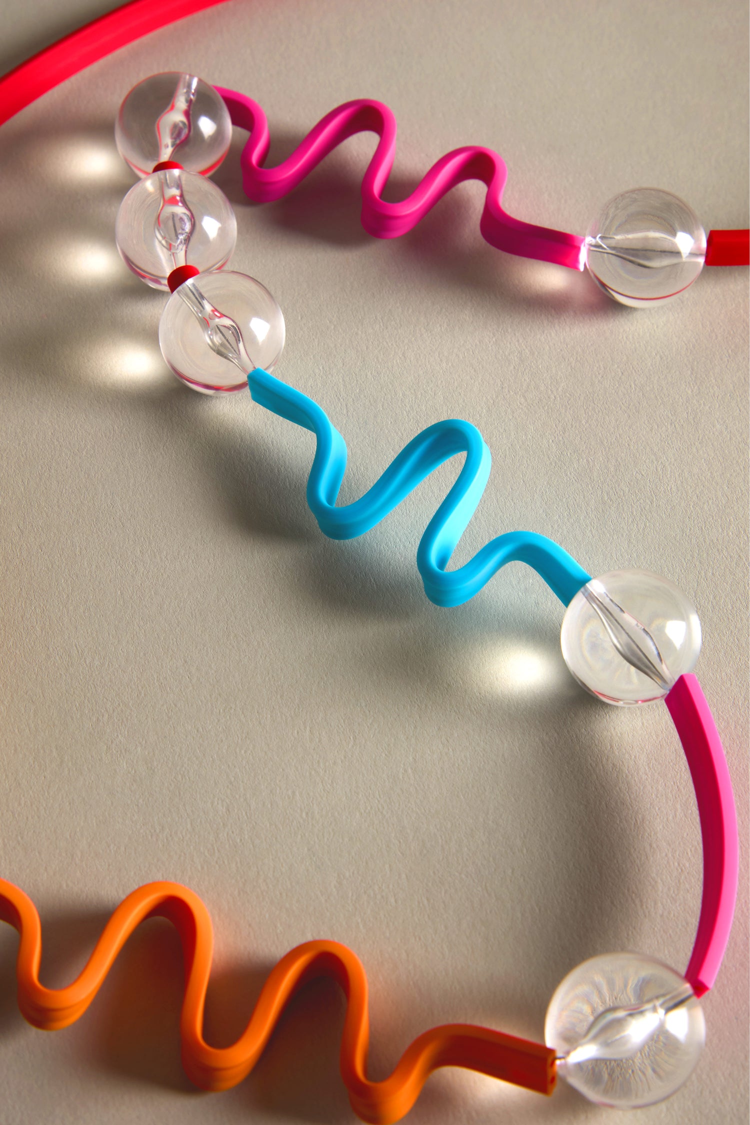 Red, blue, orange wavy tubes mingle with clear spheres on a neutral surface, highlighting Samuel Coraux's vibrant style inspired by his Squiggle And Glass Necklace.