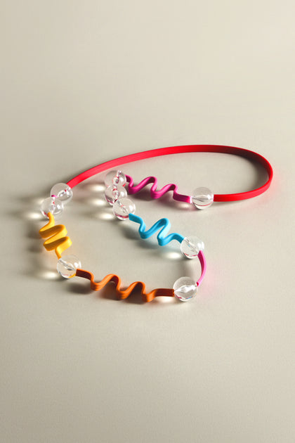 Discover the Squiggle And Glass Necklace by Samuel Coraux, showcasing bold colors with wavy orange and blue sections, clear beads, and a vibrant red cord, all set against a neutral backdrop.