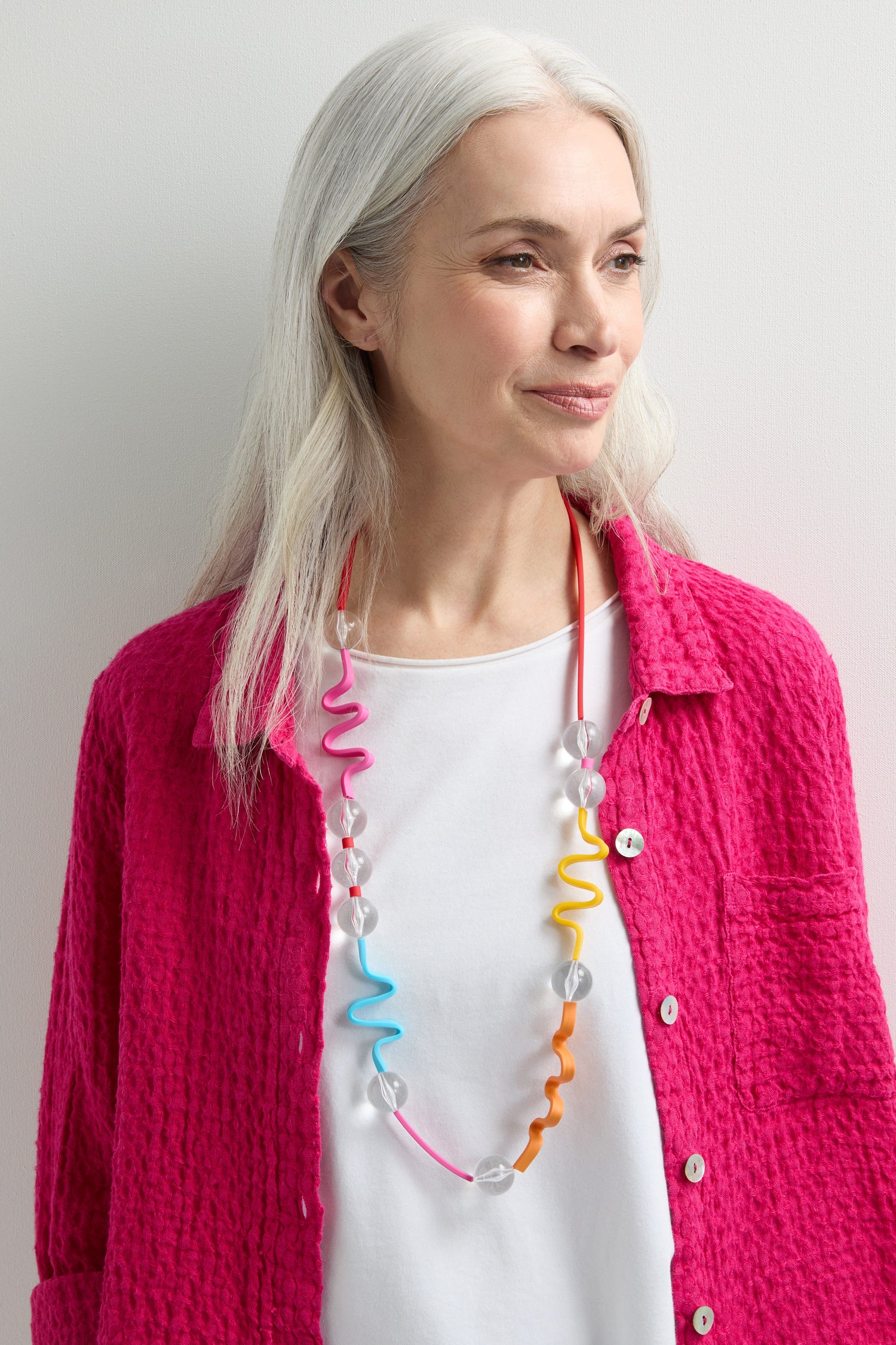 A woman with long gray hair wears a bright pink textured jacket over a white shirt, accessorized with a bold Squiggle and Glass Necklace.