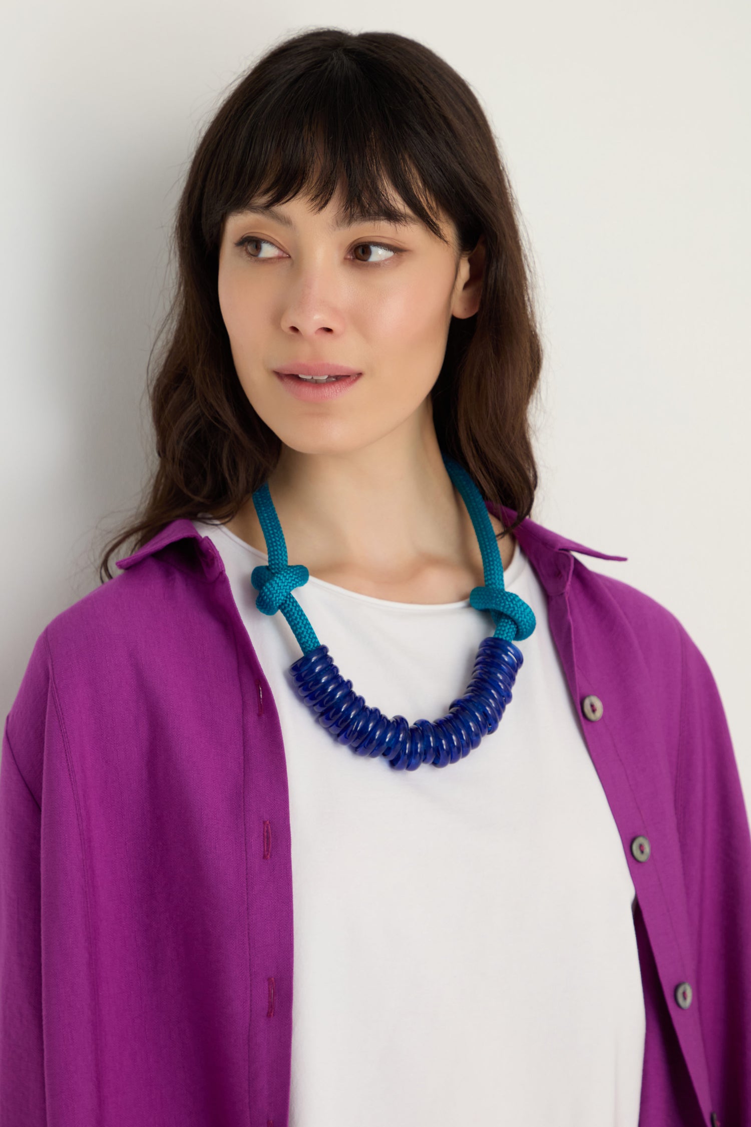 A woman wearing a bright purple shirt over a white top, accessorized with the Murano Glass Rings Necklace, looks slightly to the side against a plain background.
