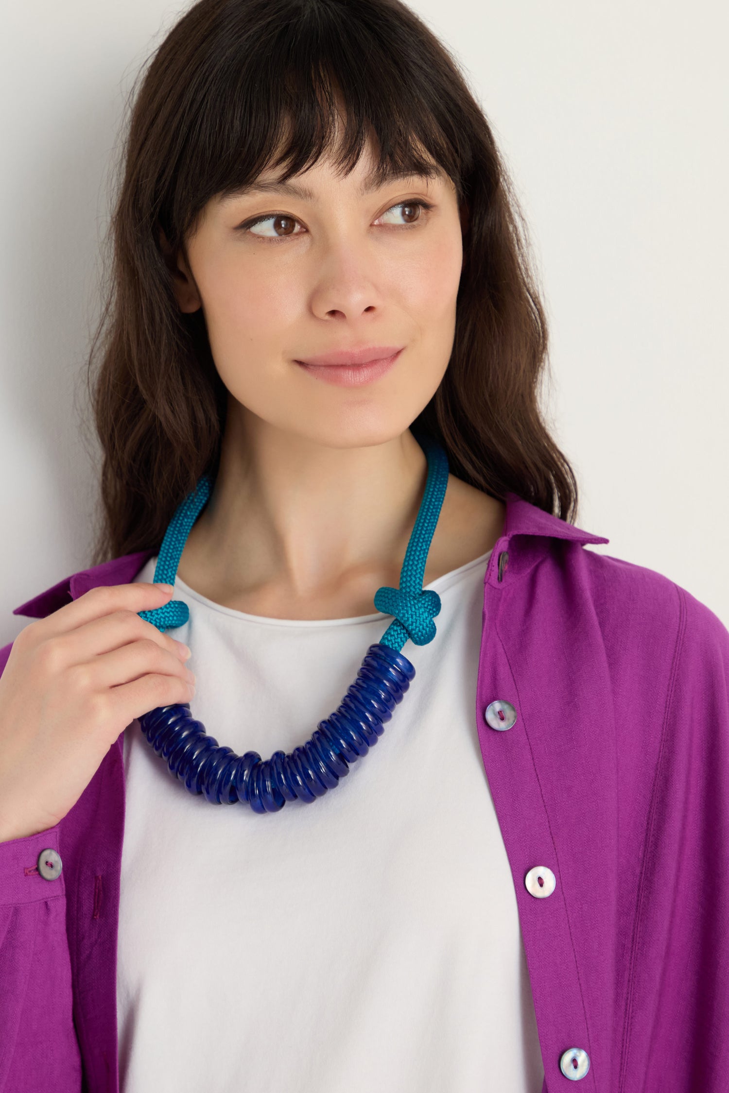 Woman with long dark hair wears a white shirt, purple button-up, and the Murano Glass Rings Necklace featuring a textured rope cord, looking slightly to the side.