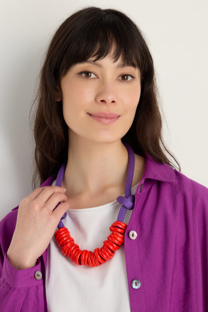 A woman in a white top and purple shirt wears the Murano Glass Rings Necklace, a chunky red and purple statement piece with a textured rope cord, standing against a plain light background.