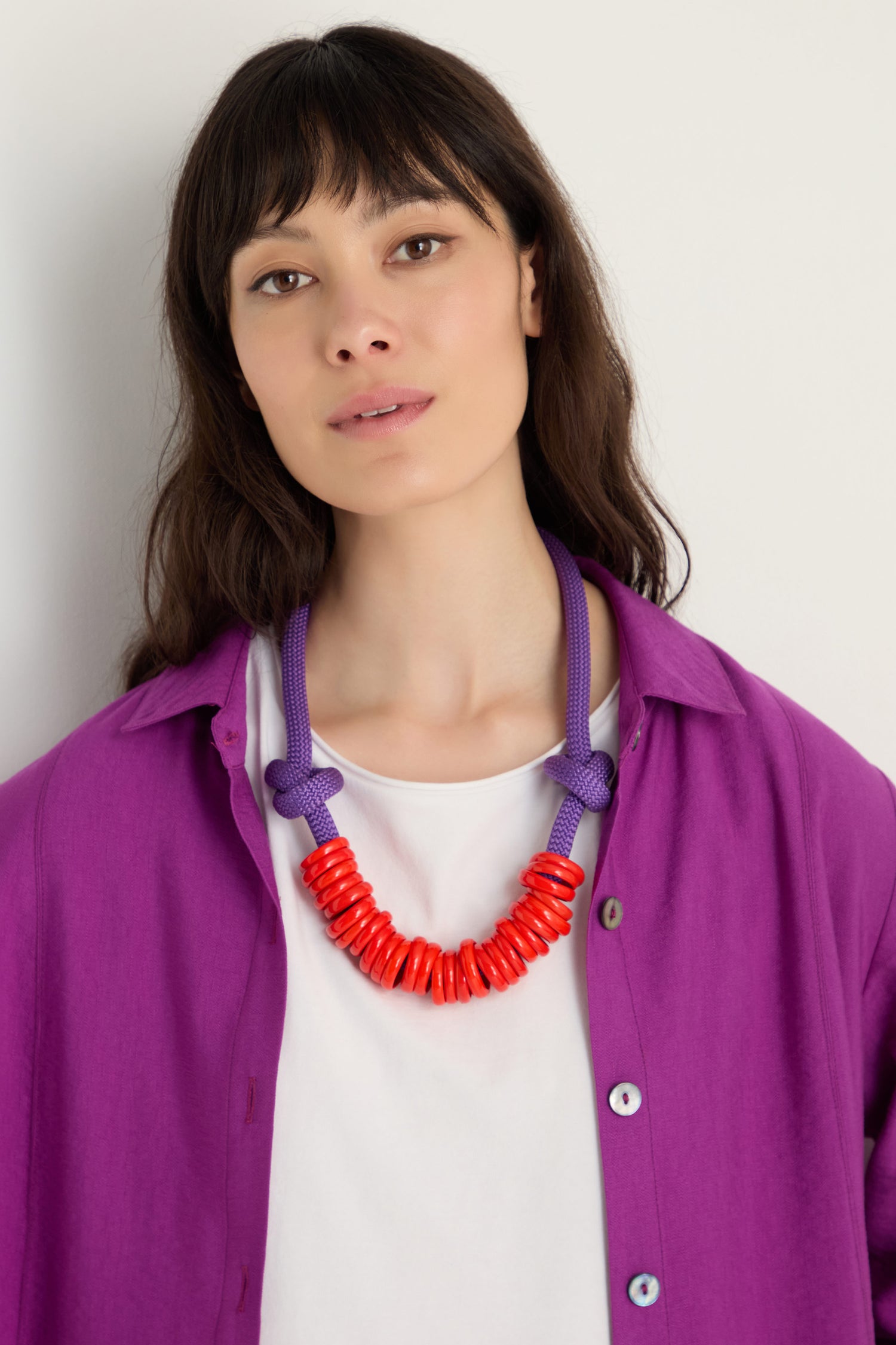 A woman stands against a plain light background, wearing a white shirt, a purple open button-up, and the Murano Glass Rings Necklace—a chunky statement piece with textured rope cord and glass rings.