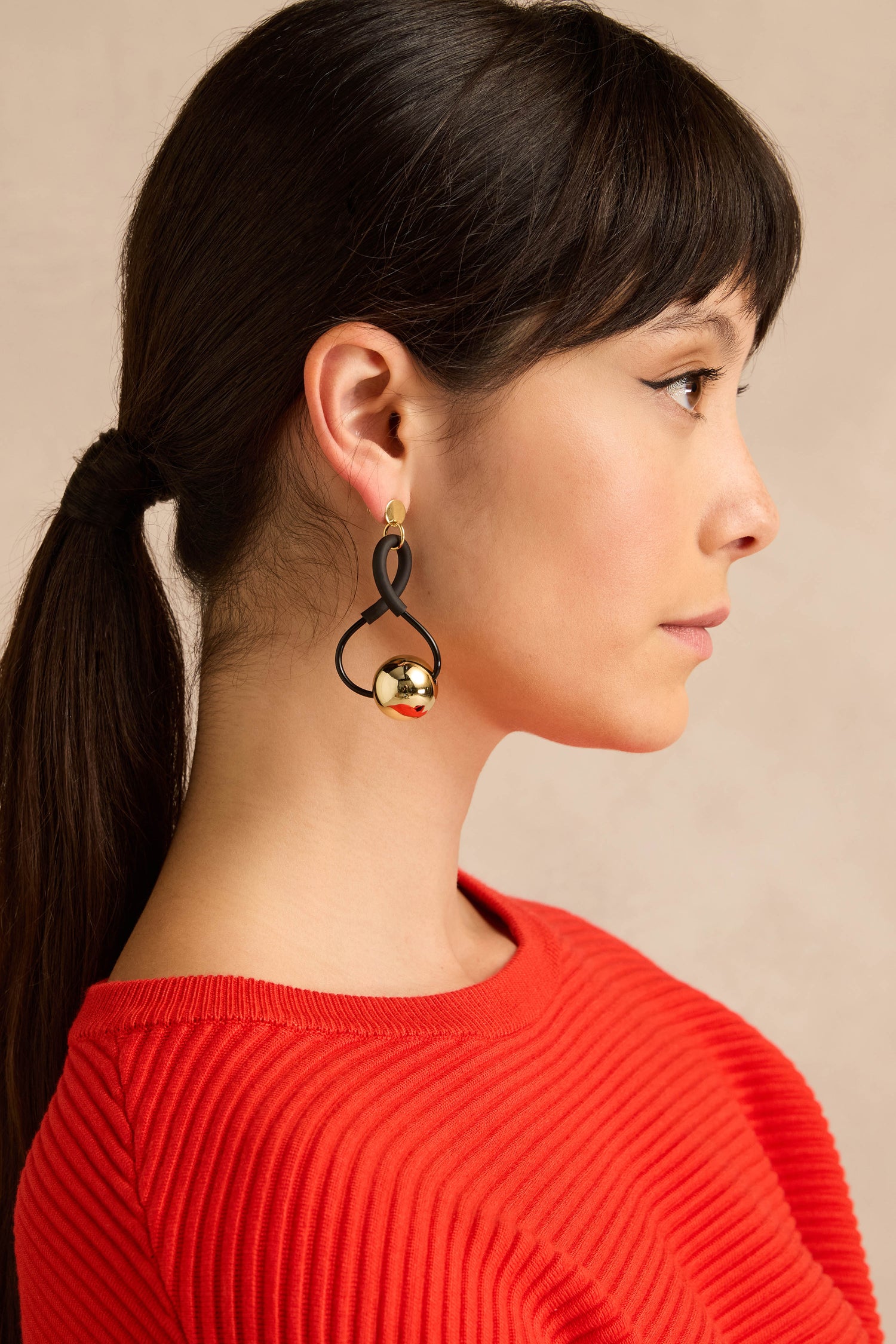A woman in profile with brown hair styled in a ponytail wears a red ribbed top and the Infinity Earrings—eye-catching black and gold earrings with a sculptural design—set against a beige background.