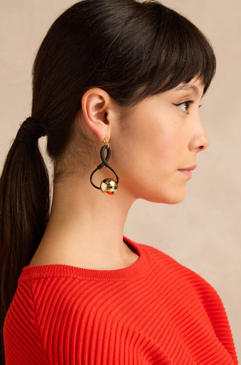 A woman in profile with brown hair styled in a ponytail wears a red ribbed top and the Infinity Earrings—eye-catching black and gold earrings with a sculptural design—set against a beige background.