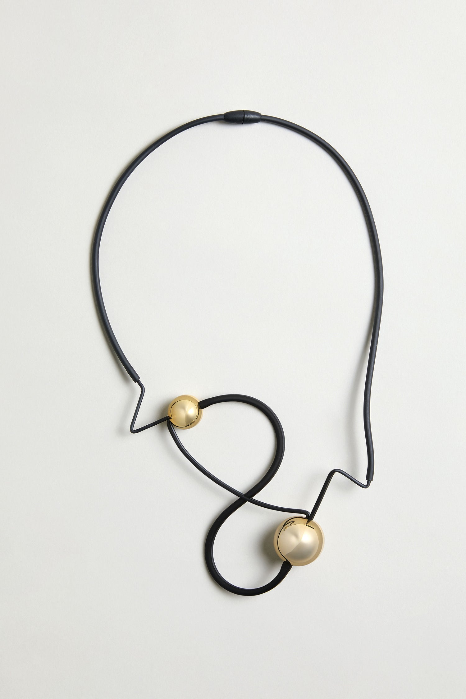 The Infinity Necklace showcases a black, abstract zigzag design with two round gold accents—one large and one small—creating a bold sculptural look, set against a simple light background.