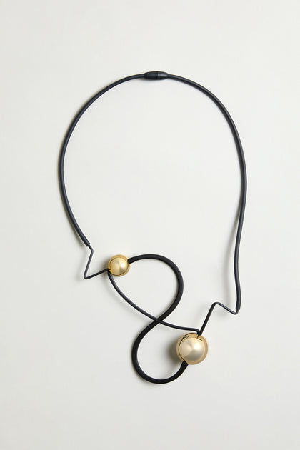 The Infinity Necklace showcases a black, abstract zigzag design with two round gold accents—one large and one small—creating a bold sculptural look, set against a simple light background.