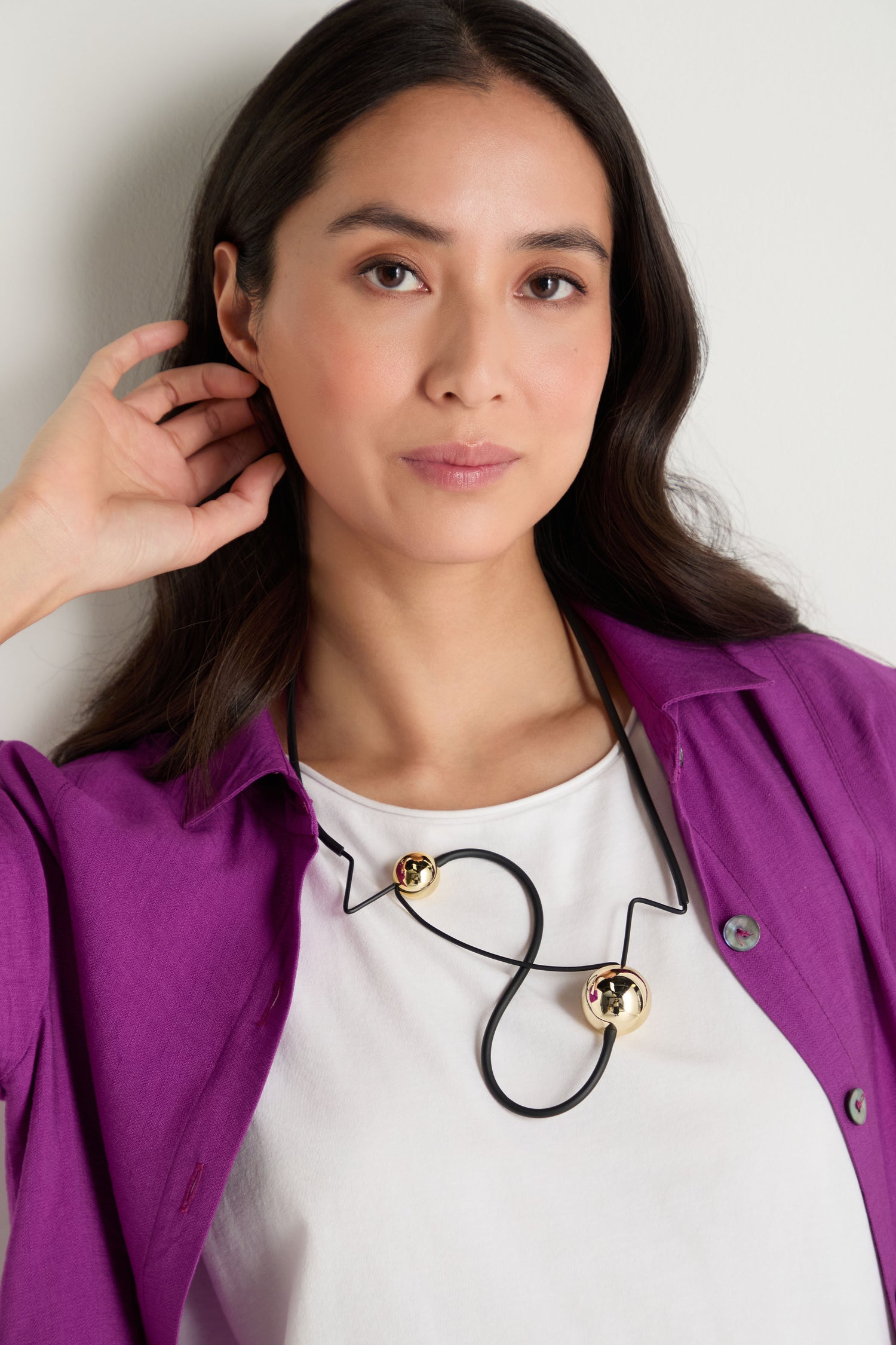 A woman with long dark hair, dressed in a white shirt and purple jacket, wears the Infinity Necklace—a bold piece of sculptural jewelry—while standing against a simple light background.