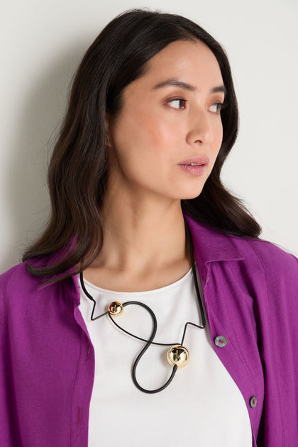 A woman wearing a white shirt, purple jacket, and the Infinity Necklace—featuring striking black sculptural design with two gold spheres—stands against a light wall, gazing to the side.