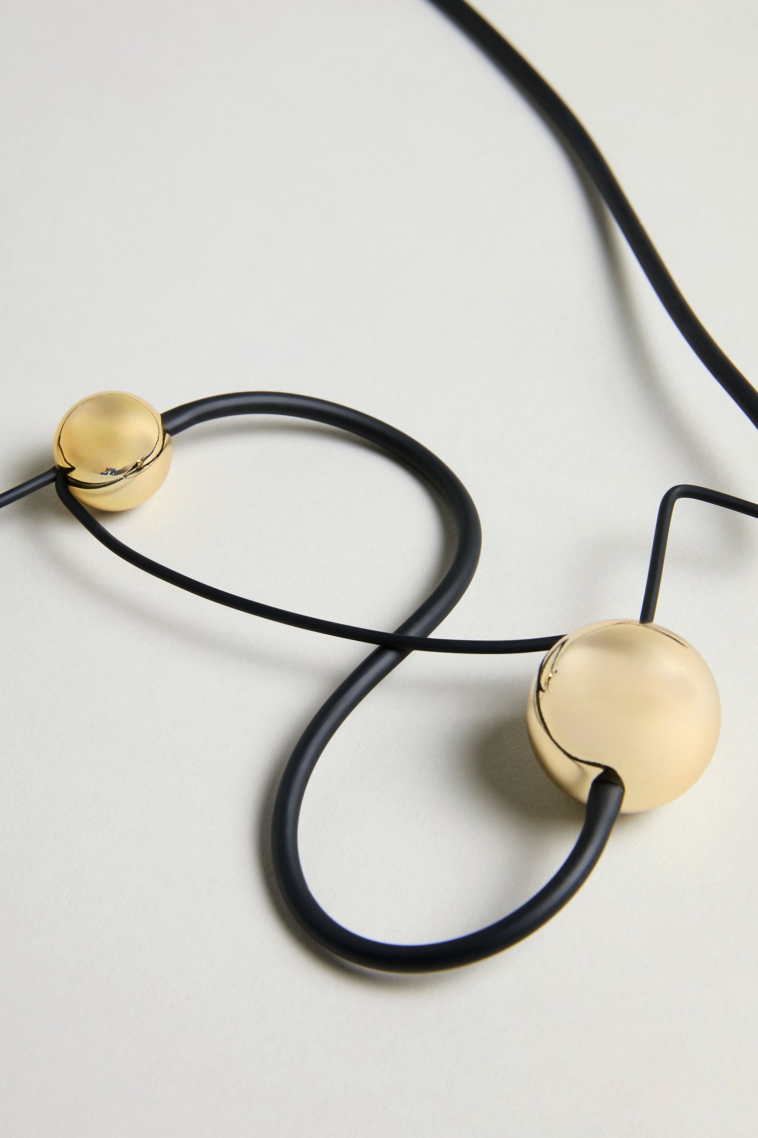 The Infinity Necklace features a black cord with two large, round gold-colored beads, displayed on a light surface—an eye-catching piece of sculptural jewelry.