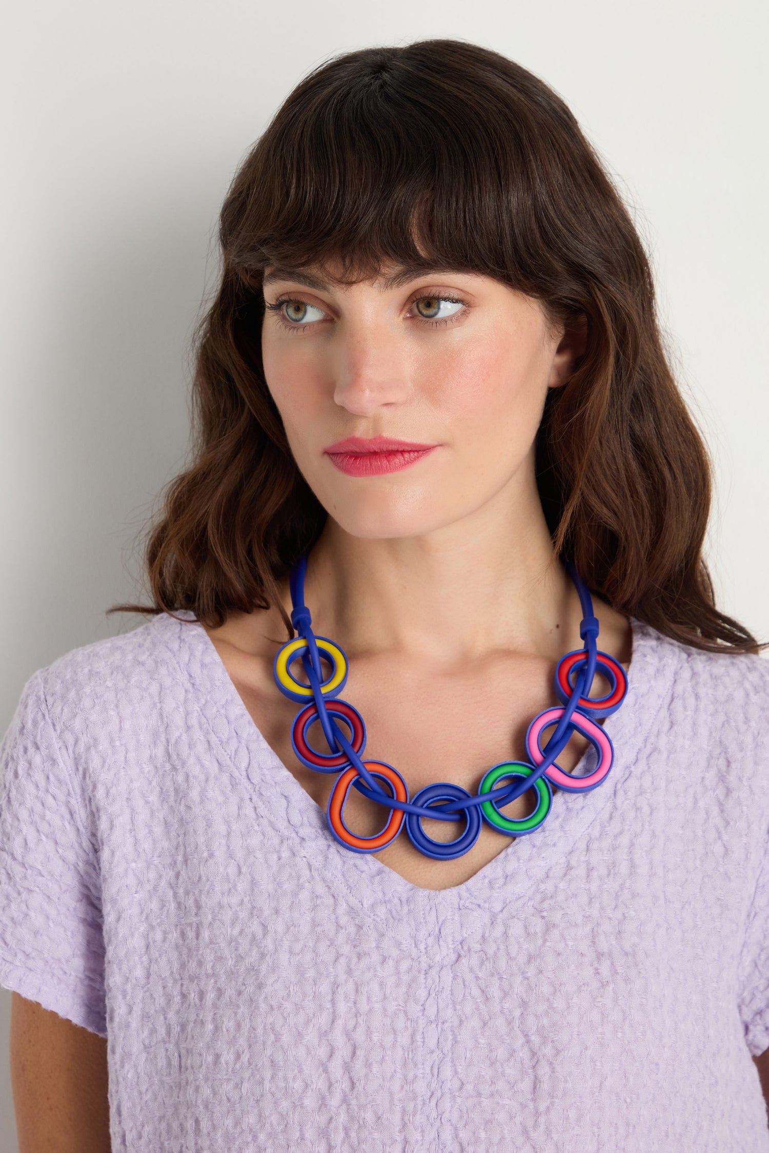 A woman with brown hair wears a textured light purple top and the Colour Pop Rings Necklace, which features an interlocking ring design, as she looks slightly to the side against a plain background.