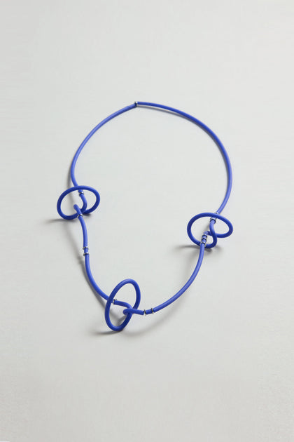 The Triple Twist Necklace is a modern blue statement piece with three interlocking circular loops, elegantly arranged on a flat, light background.