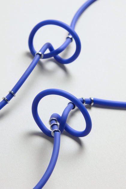The Triple Twist Necklace features flexible blue tubes looped through bold blue rings and secured with metallic clamps, creating a striking, modern statement piece displayed on a neutral light background.