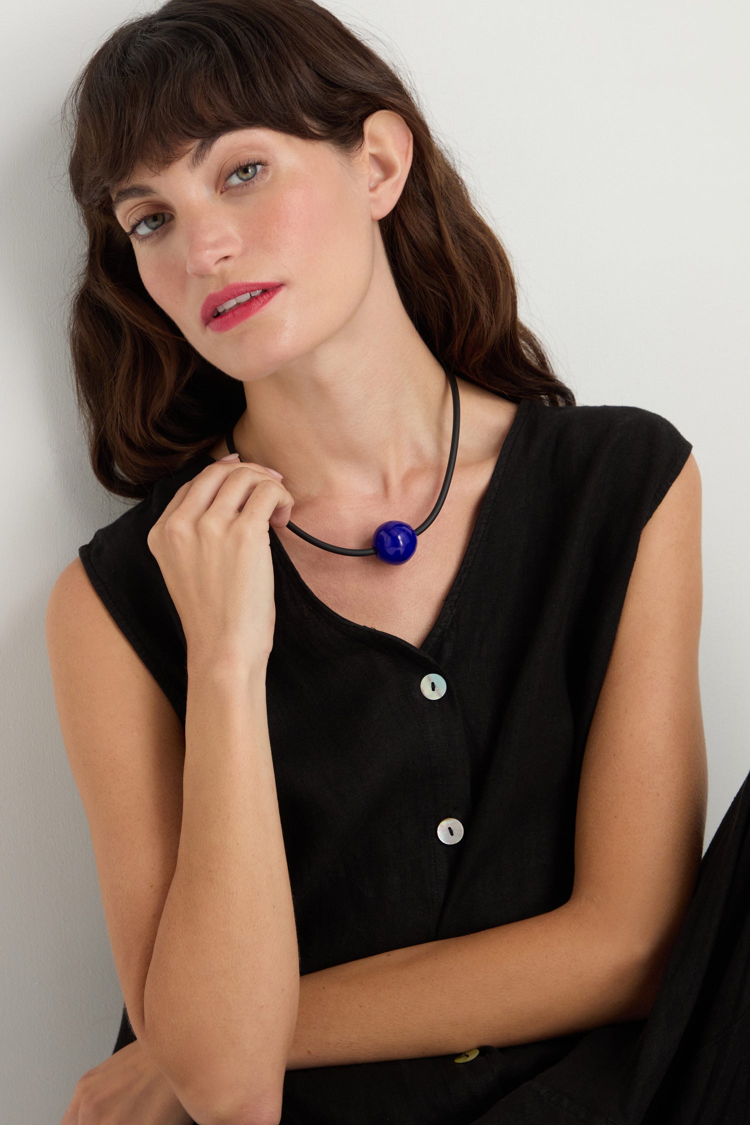 A woman with dark hair, dressed in a sleeveless black top, wears the Murano Bead Necklace—a minimalist accessory with a blue Murano bead—while sitting against a white wall.