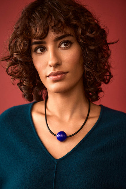 A woman with curly brown hair, dressed in a teal top and wearing the Murano Bead Necklace—a handmade black necklace from Paris featuring a large blue bead—stands in front of a plain red background.