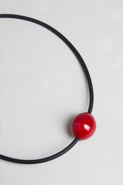 The Murano Bead Necklace is a handmade minimalist piece from Paris with a black circular design and a glossy red Murano bead near the bottom, shown on a plain white background.