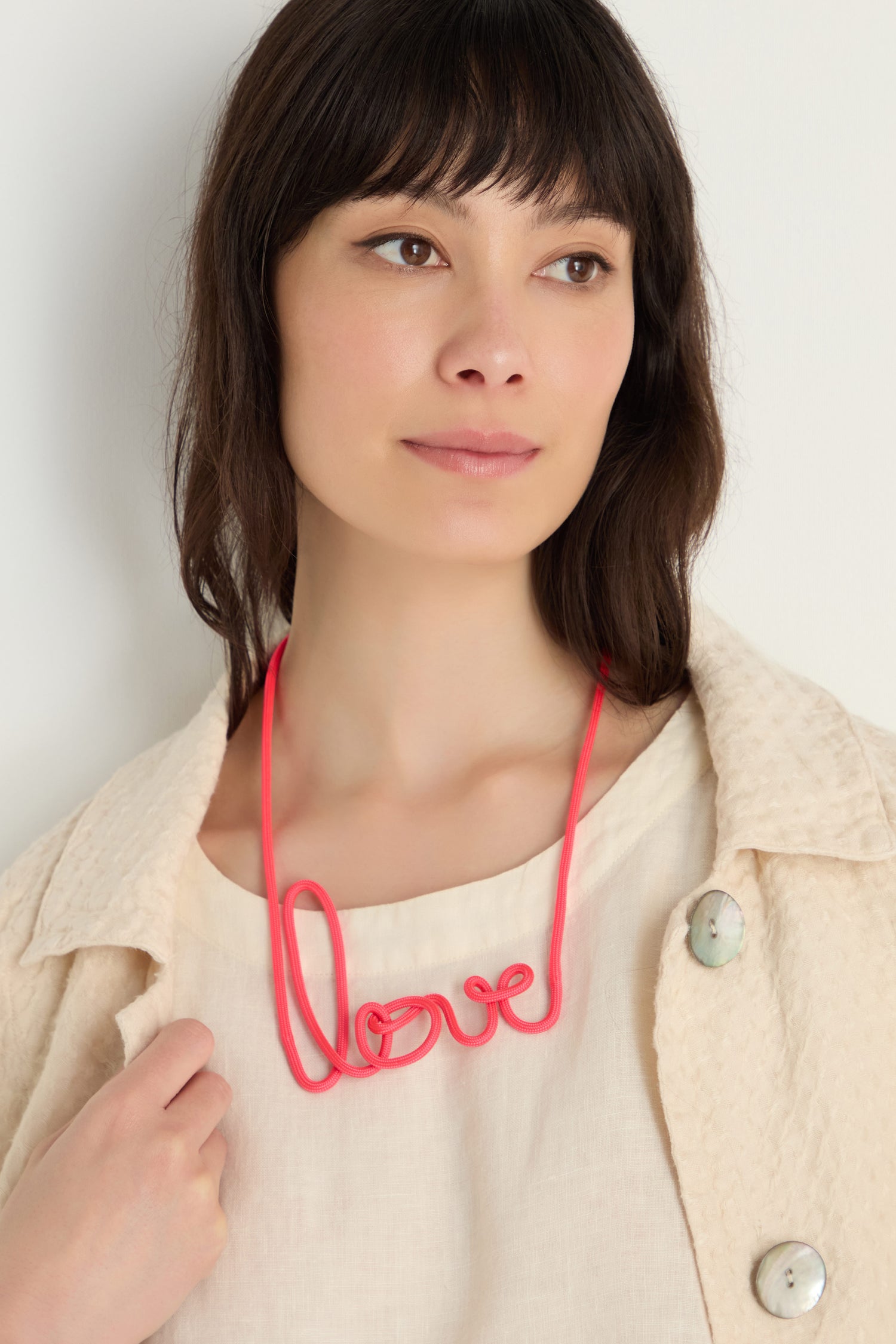 Woman with straight brown hair wears a cream textured jacket and the Love Necklace, a red graphic accessory handmade to spell "love.