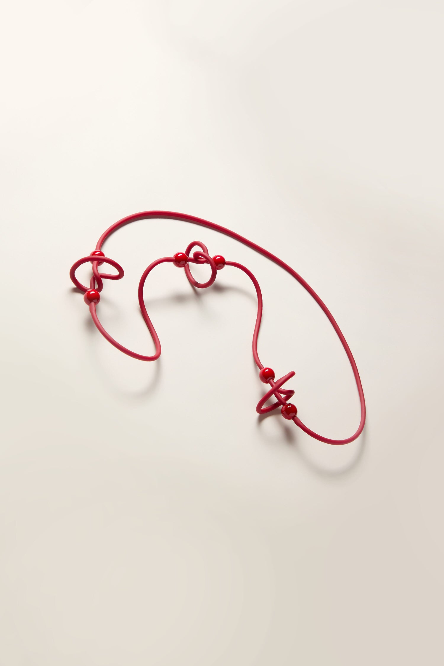 The Trio Squiggle Necklace by Samuel Coraux, featuring a red wire-like structure with three large knots and several smaller beads, is laid out against a plain light background, showcasing its abstract design.