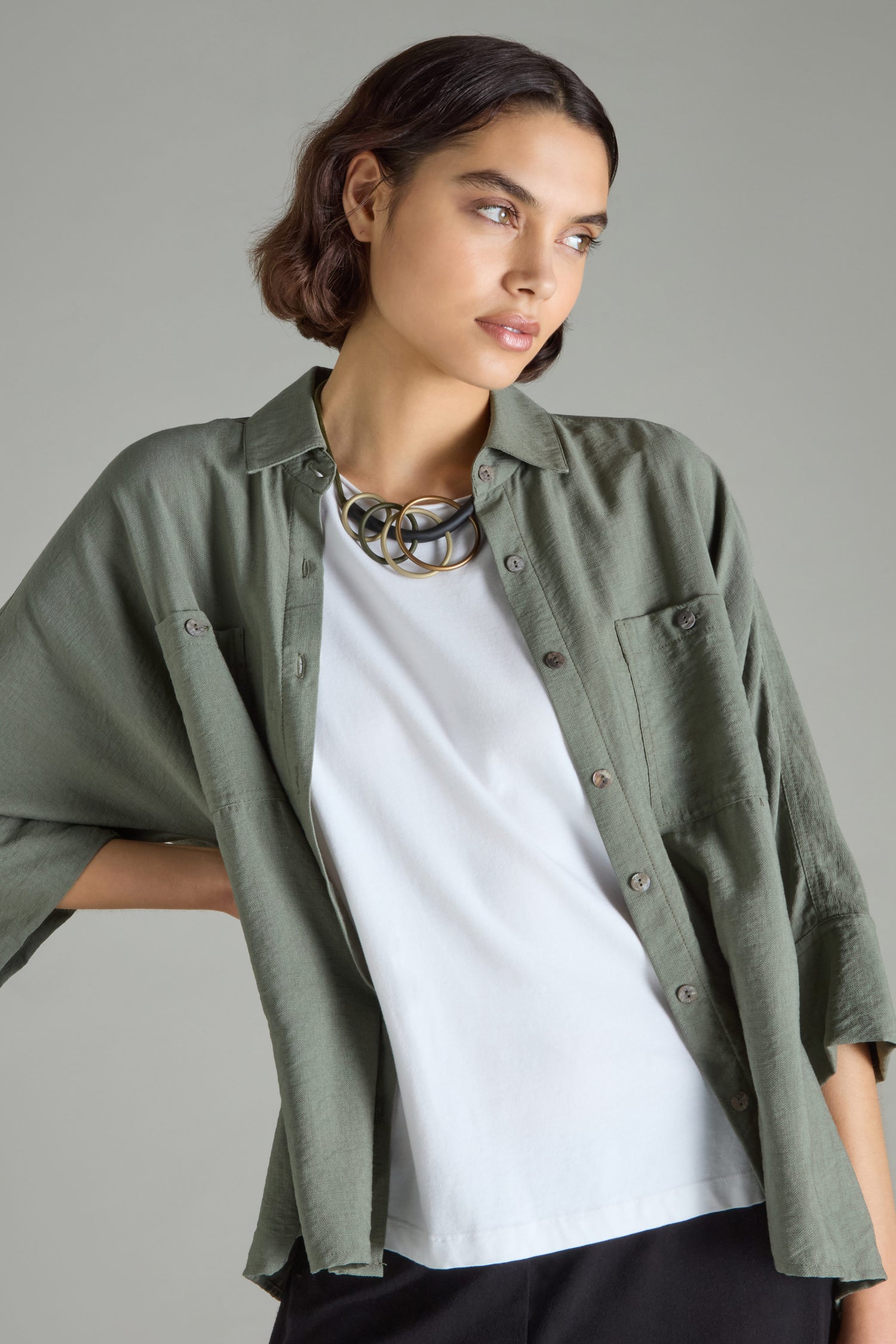 A person donning a green button-up shirt layered over a white top and an oversized Quadruple Circles Necklace by Samuel Coraux, radiating modern elegance as they gaze to the side against a neutral backdrop.