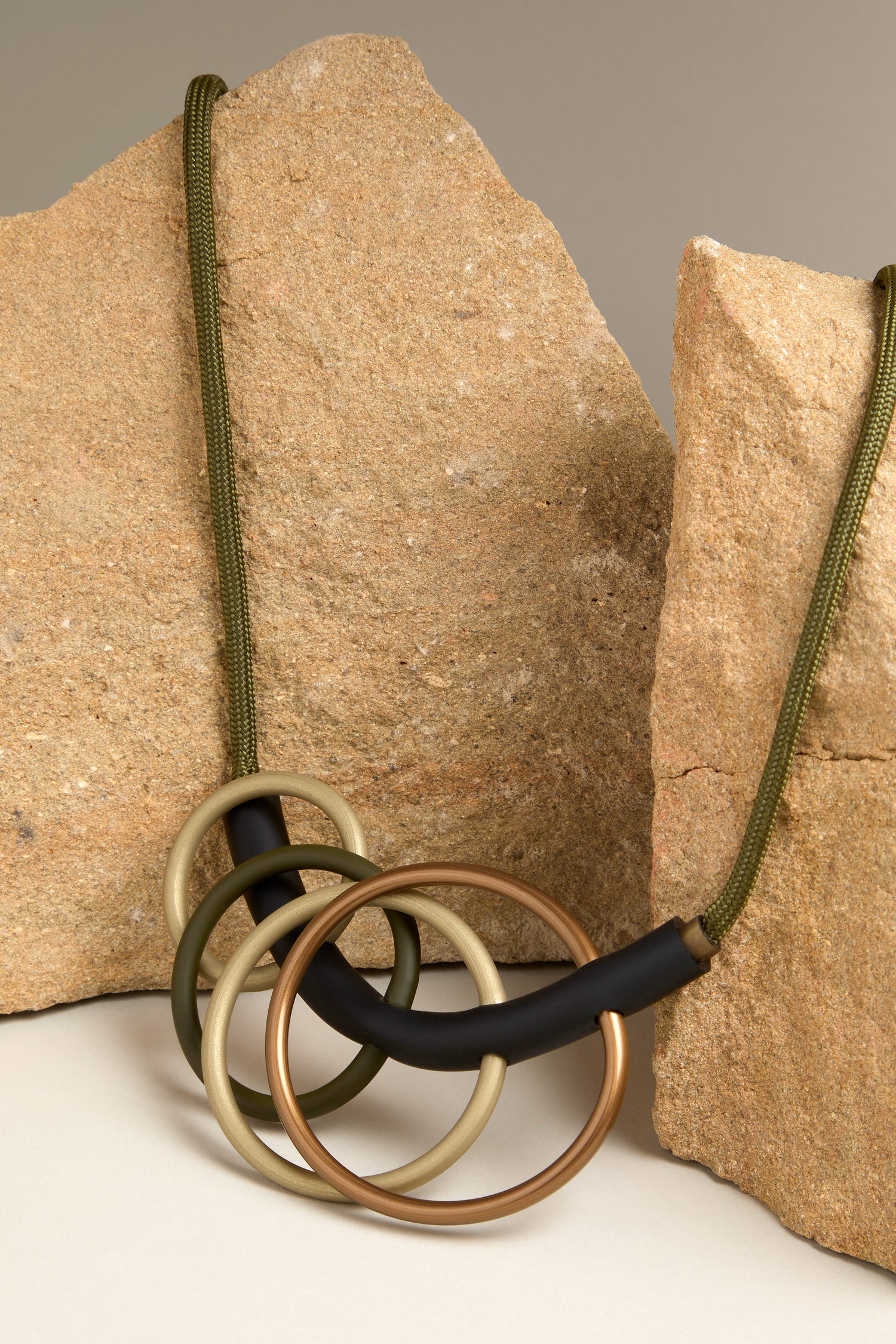 The Quadruple Circles Necklace exudes modern elegance with its interlocking metal rings in diverse finishes, artistically arranged on a green cord. Presented against rough-textured stone slabs, it epitomizes a contemporary aesthetic that is both striking and sophisticated.