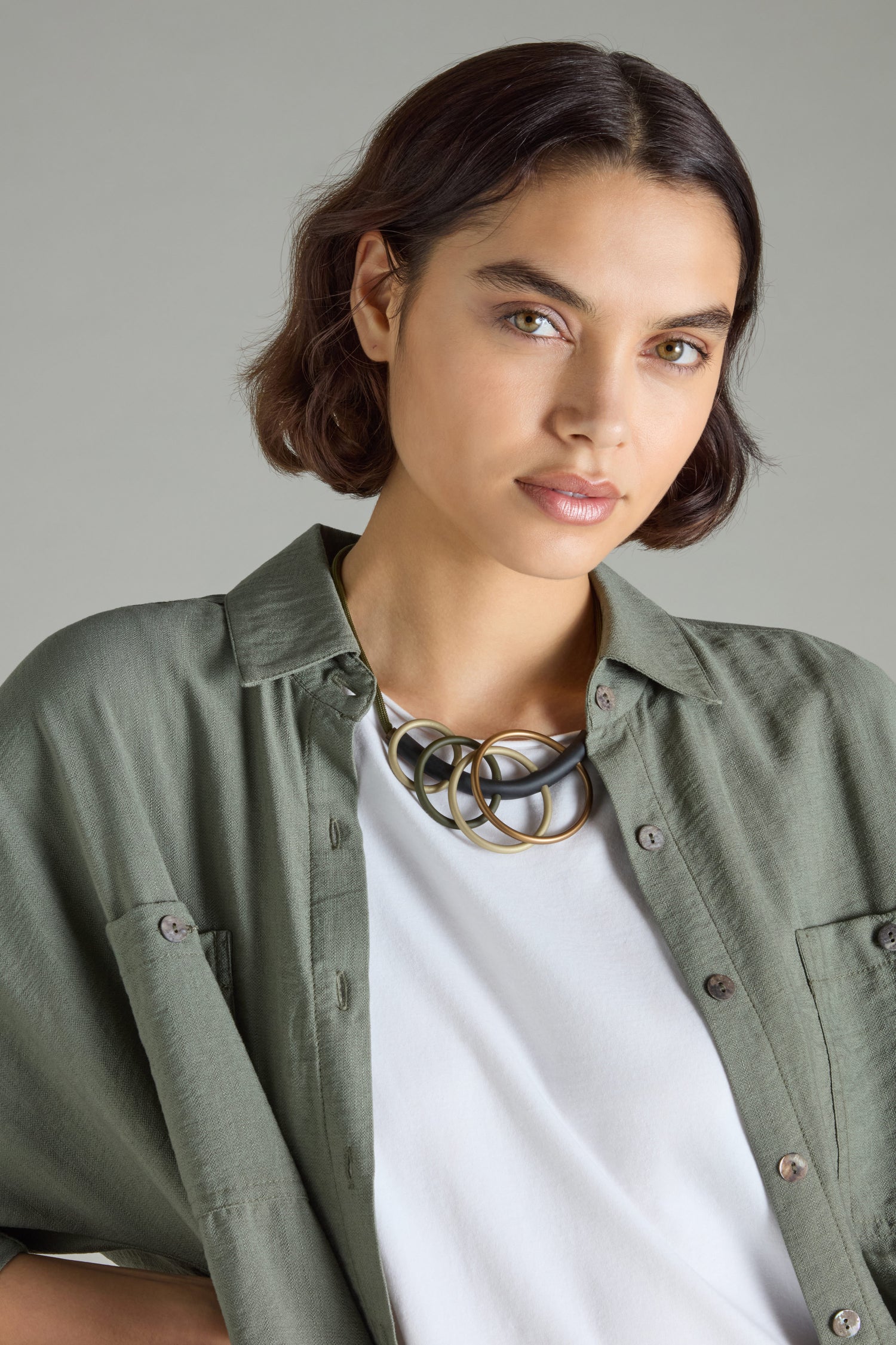 A person with short brown hair is wearing a green button-up shirt over a white top and beautifully highlights the modern elegance of the Quadruple Circles Necklace by Samuel Coraux. They appear to be looking directly at the camera against a neutral background.