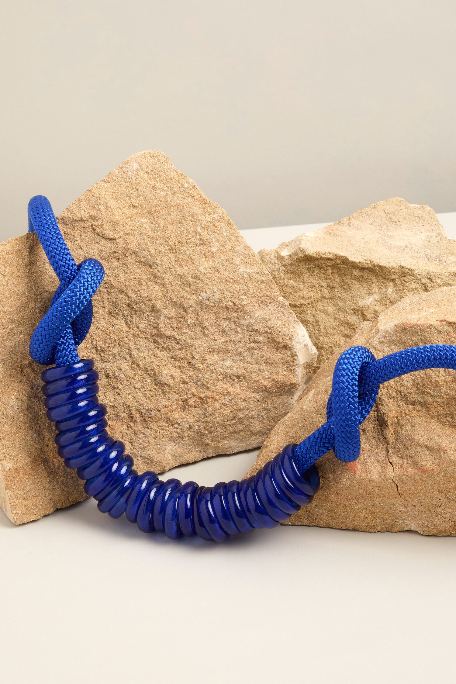 A blue coiled rope tied in knots rests on two large rocks, reminiscent of the Murano Glass Rings Necklace by Samuel Coraux.