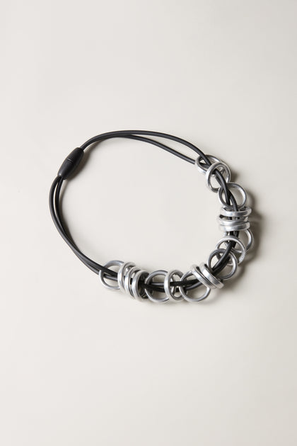 The Black and Silver Metal Rings Necklace features multiple large, interlocking silver metal rings clustered on one side of a black cord, set against a light background. Inspired by the bold designs of Samuel Coraux Jewelry.