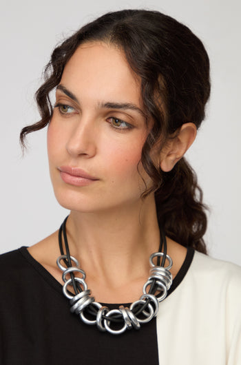 A woman with wavy brown hair in a black and white top models the Black and Silver Metal Rings Necklace by Samuel Coraux, a contemporary statement piece, as she looks slightly to the side against a plain background.