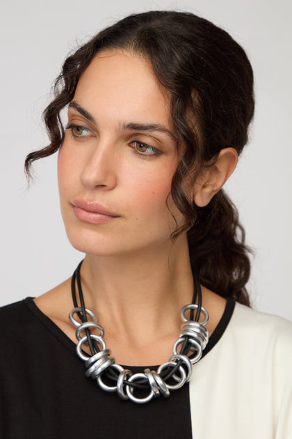 A woman with wavy brown hair in a black and white top models the Black and Silver Metal Rings Necklace by Samuel Coraux, a contemporary statement piece, as she looks slightly to the side against a plain background.