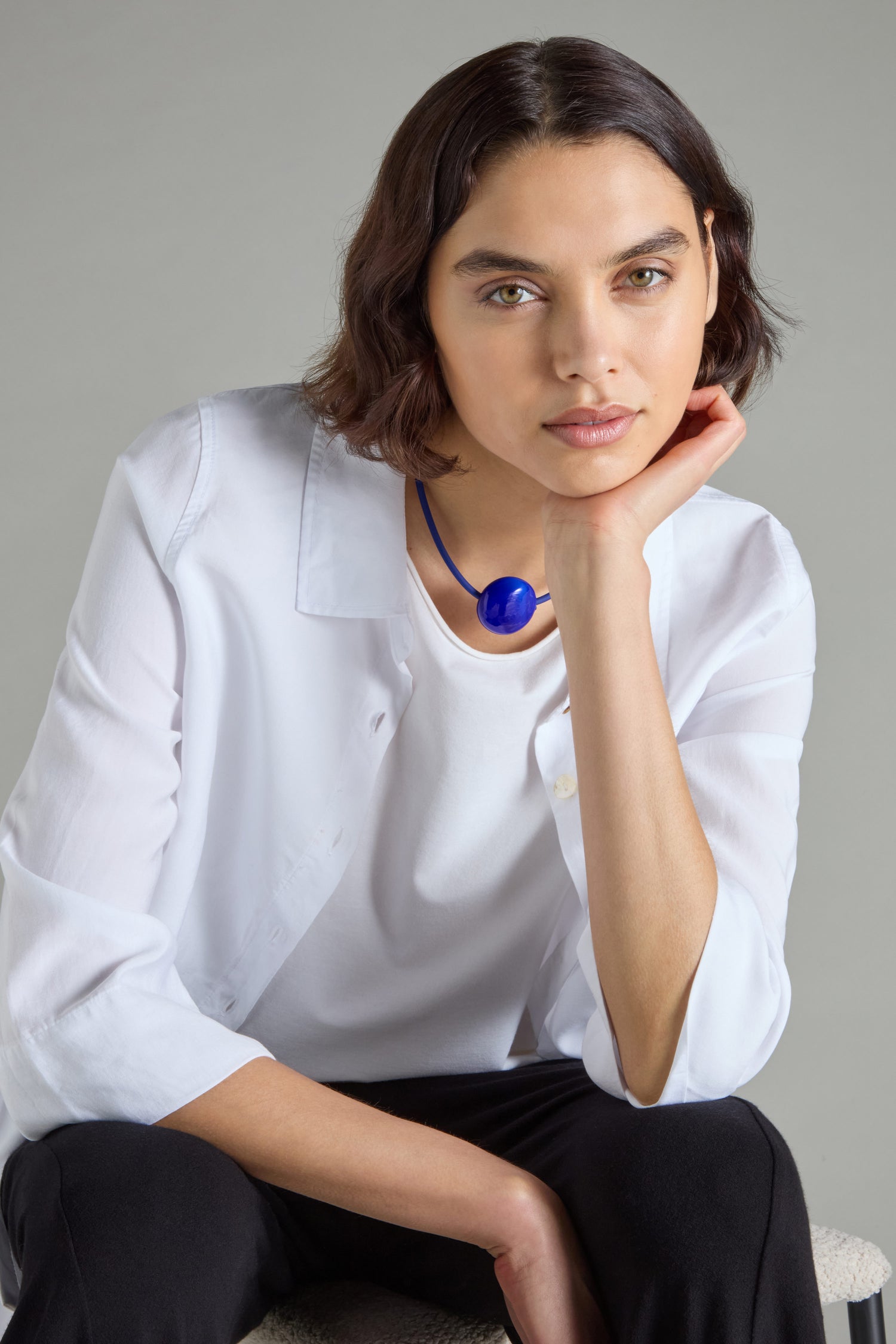 A person with short brown hair, wearing a white shirt and a striking blue Murano Glass Pebble Necklace, handcrafted in France by Samuel Coraux, sits with their chin resting on one hand, looking at the camera.