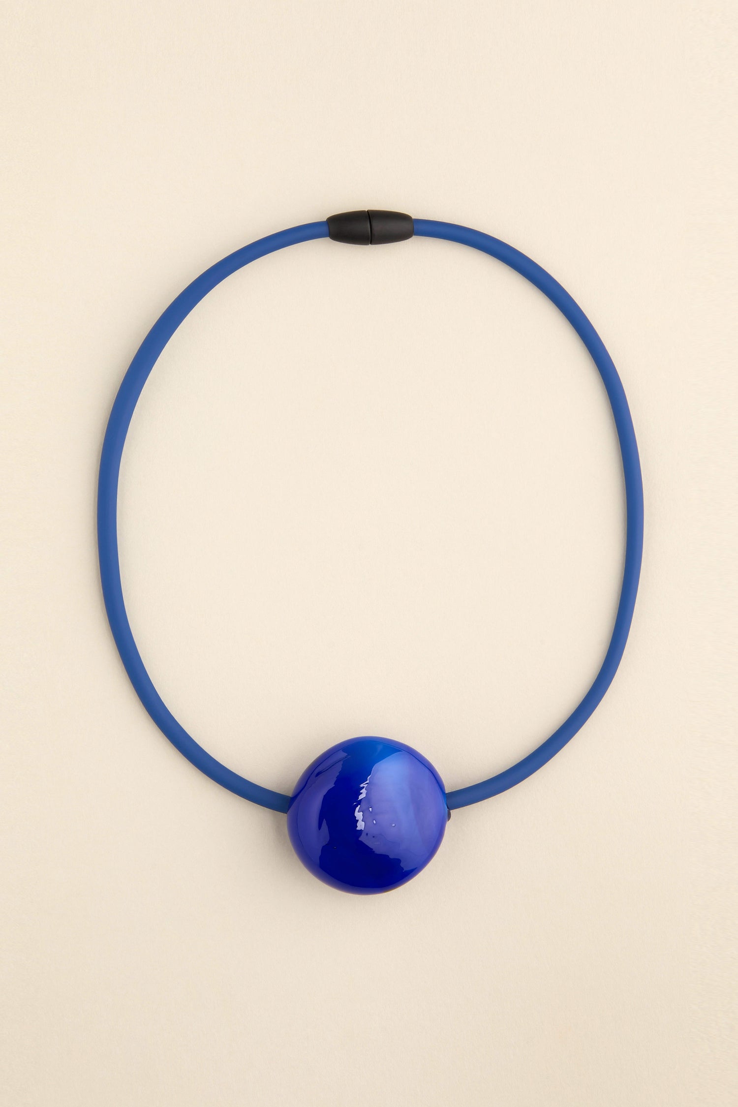 The Murano Glass Pebble Necklace, designed by Samuel Coraux, is an eye-catching accessory crafted in France. It showcases a prominent spherical pendant strung on a coordinating blue cord and fastened with a stylish black clasp.