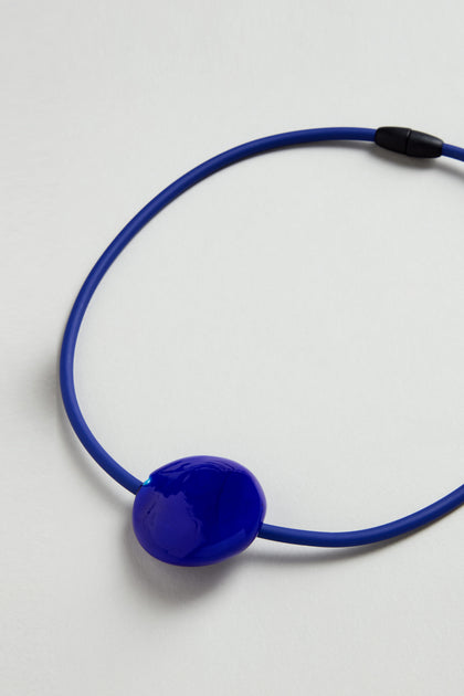 The Murano Glass Pebble Necklace features a blue cord, glossy irregular blue pendant, and black clasp, all displayed on a light background.