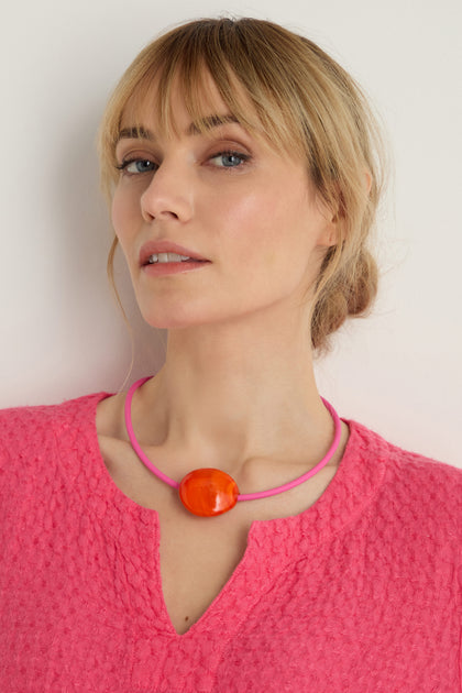 A woman with blonde hair in a loose bun wears a textured pink top and the Murano Glass Pebble Necklace featuring a large orange pendant, striking a pose against a light background.