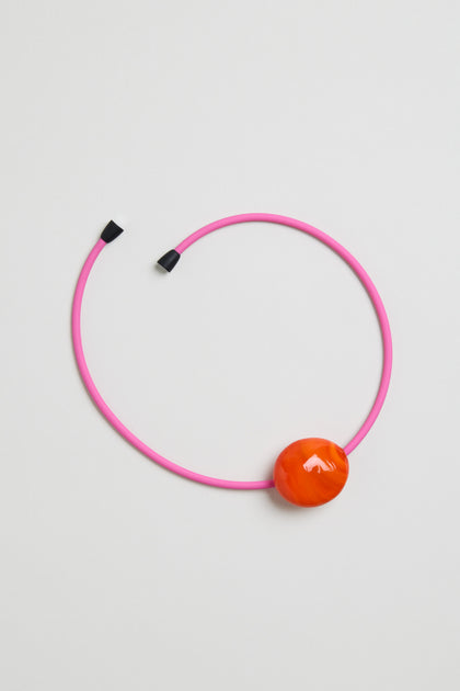 The Murano Glass Pebble Necklace features a pink choker with black tips and a large round orange bead, shown on a white background. This bold, eye-catching design evokes the classic pebble necklace style.