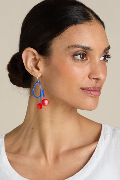 A woman with dark hair tied back wears a white top and blue Symphony Earrings featuring red spheres. She faces slightly to the right against a neutral background, her playful and bold statement adornment standing out vividly.